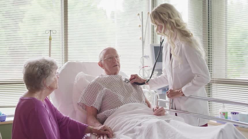 Female doctor listens to male patients heart with a stethoscope as his wife sits beside him and holds his hand