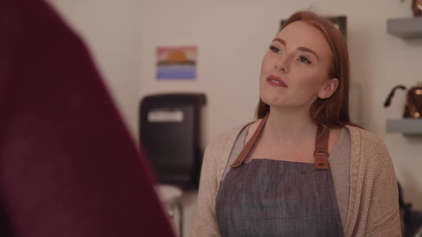 Woman with red hair fair skin and a blue apron talks to person and moves her head up and down as a way of agreeing in a cafe