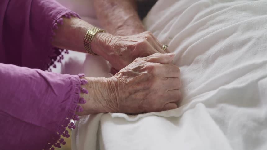 An elderly woman sitting beside a bed grasps the hand of an elderly man lying within the bed