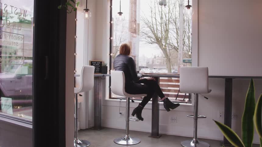 Woman with red hair a black leather jacket sits at a table and looks out the cafe windows