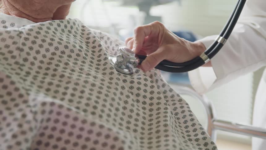 A doctor uses a stethoscope to listen to the chest of an elderly man lying in a hospital bed