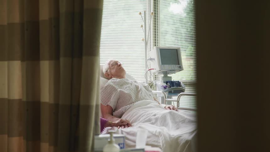 An elderly patient rests in a hospital bed as an elderly woman sitting beside the bed holds his hand