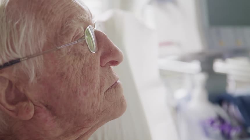 An elderly man sits in a hospital bed propped up with a pillow behind his head