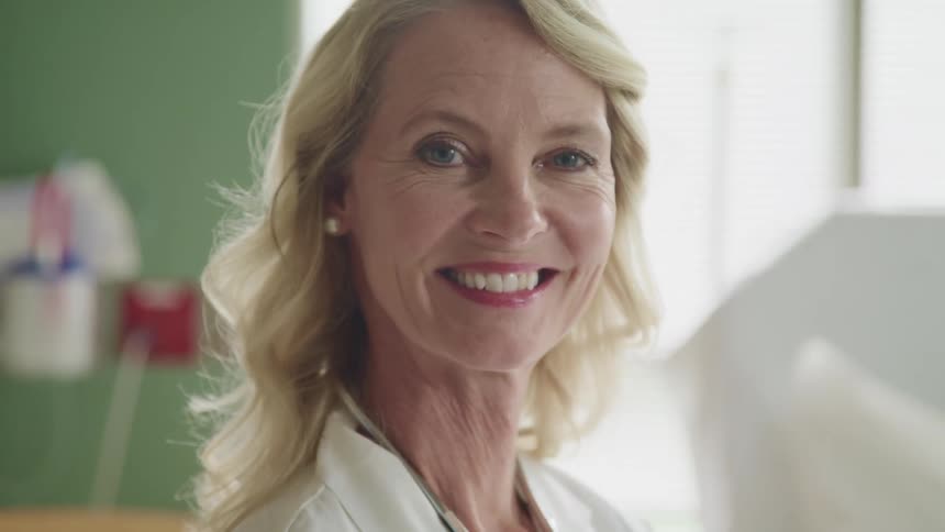 A woman with a lab coat and stethoscope turning around and smiling with a window in the background