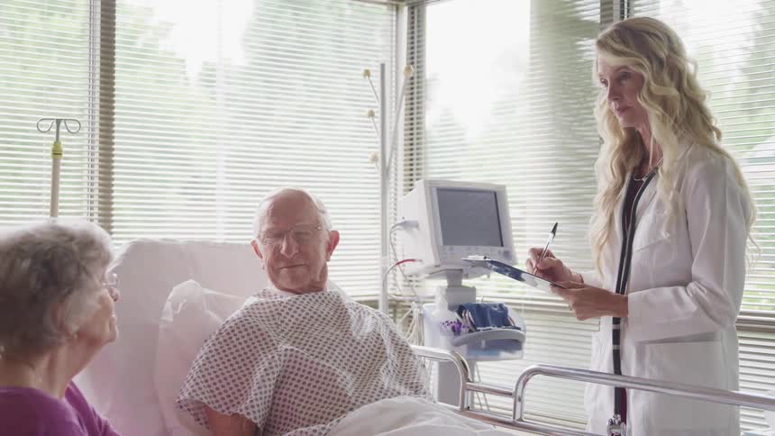 A female doctor stands beside an elderly male patient in his bed with his wife sitting beside him
