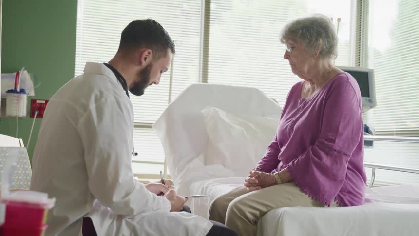 A male doctor sits beside an elderly female patient on a hospital bed