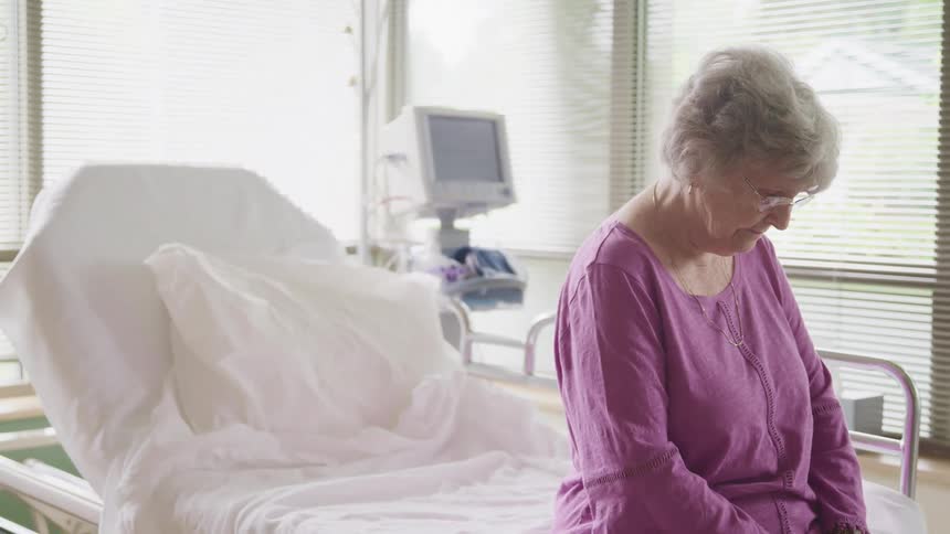 An elderly woman sits on an empty hospital bed with her head hung down low