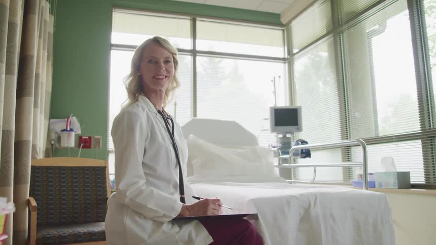Female doctor wearing lab coat and stethoscope sits next to hospital bed smiling at the camera