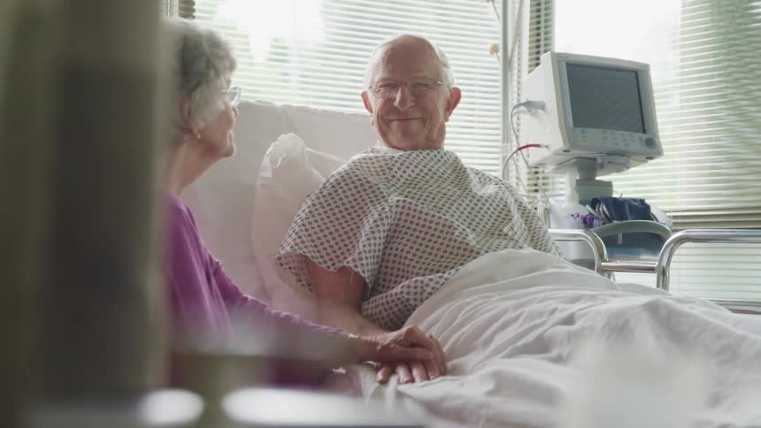 An elderly woman sits beside her husband in a hospital bed
