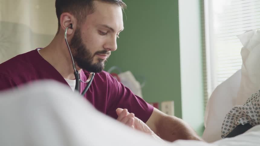A man with a stethoscope and scrubs tending to a patient laying in a hospital bed
