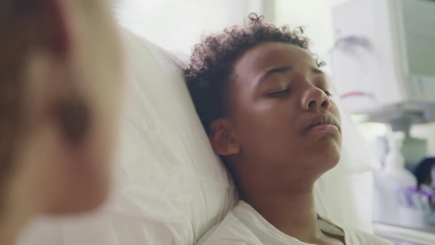 A mother sits next to her son as he sleeps in a hospital bed