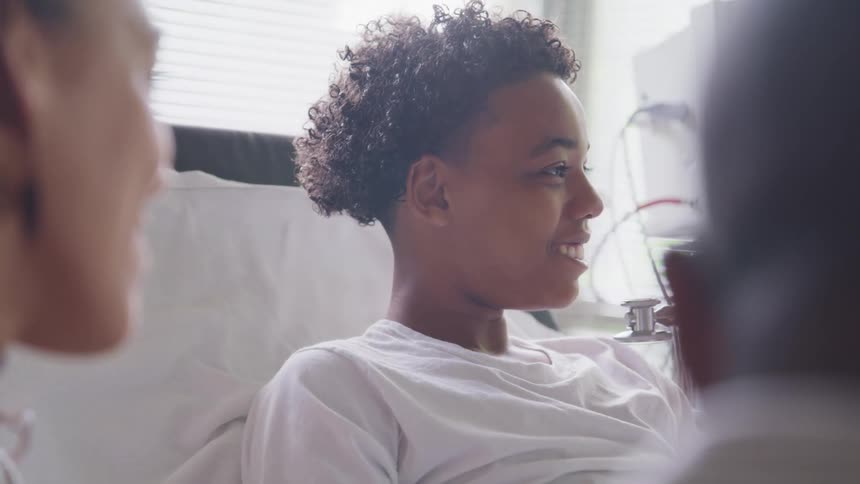 A boy smiles as he rests in a hospital bed while a doctor examines him with a stethoscope