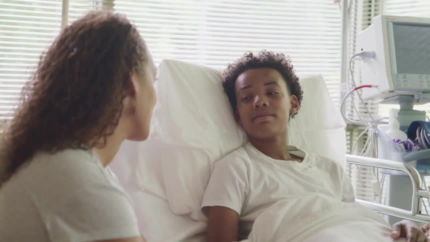 A young boy lies in a hospital bed and looks at his mother seated next to him