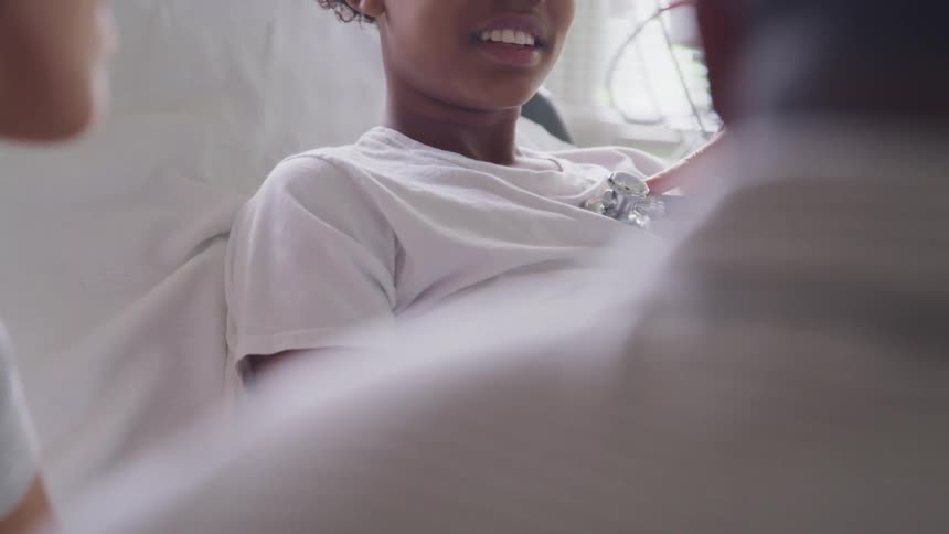Patient sits in hospital bed smiling while doctor checks heart as parents wait by bedside