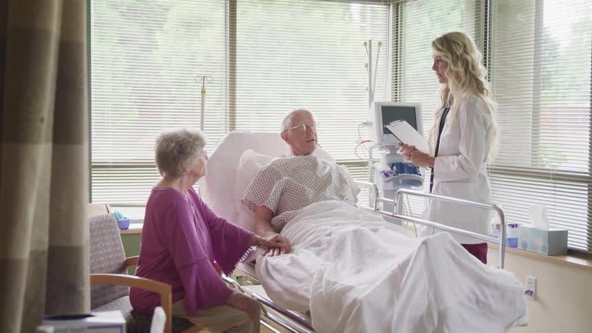 An elderly woman holds her husbands hand as he lies in a hospital bed and they talk with a doctor