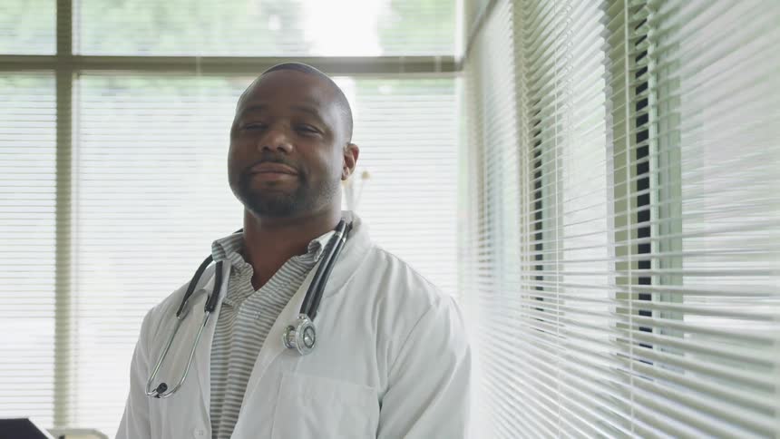Black doctor with a lab coat and a stethoscope around his neck smiles slightly and faces the camera with eye contact in the clinic