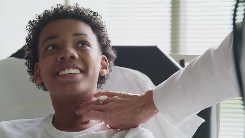 A boy in a clinic getting his pulse read by a doctor with a window and blinds in the background
