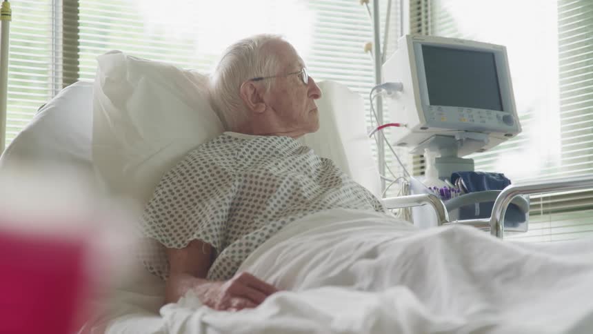 Elderly man lays on a hospital bed looking out the window surrounded by medical equipment