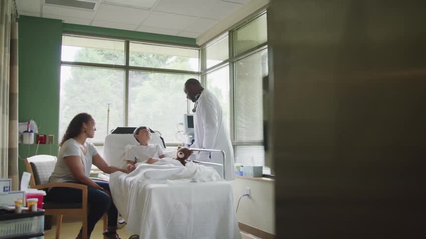 A doctor talks with a boy in a hospital bed as his mother holds his hand