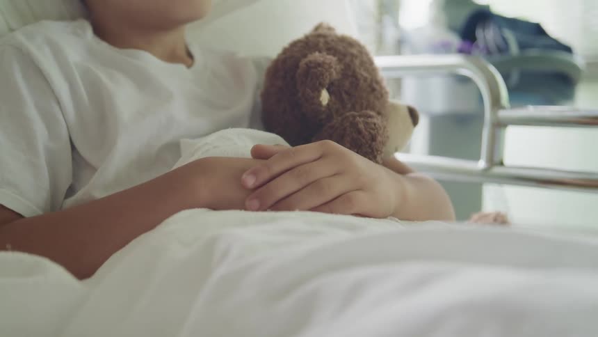 A person laying in a hospital bed with a blanket holding a teddy bear