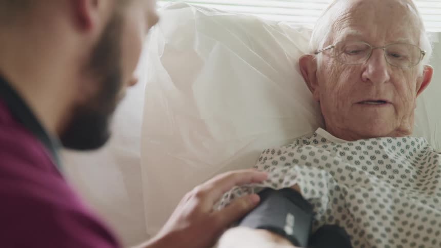 An elderly patient is being prepared for a blood pressure check by a young male nurse in a hospital