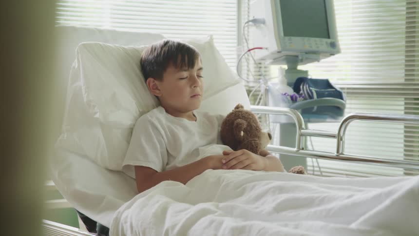 A young boy lays in a hospital bed holding a teddy bear