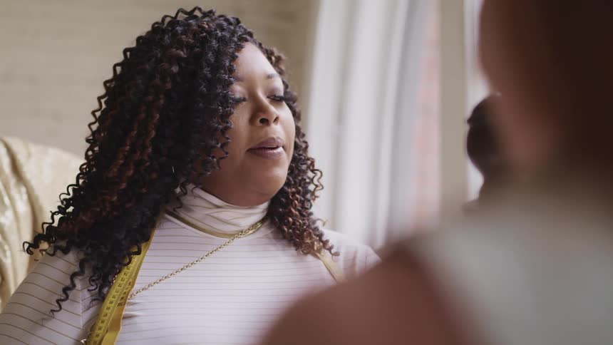A black woman with a measuring tape hanging from her shoulder happily speaks with another woman as they stand together near a window
