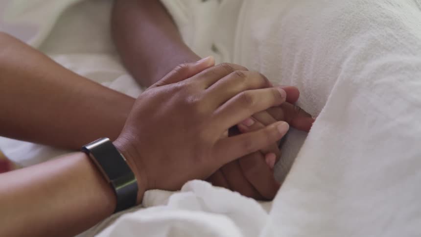 Person clasps hand of patient lying in hospital bed