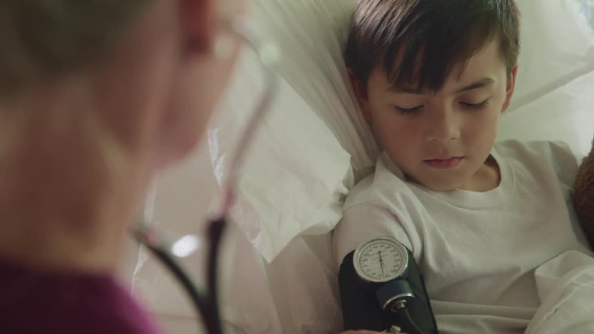 Female nurse uses a stethoscope and blood pressure cuff to check male childs blood pressure while male child watches as he is lying in a hospital bed and holding onto a teddy bear