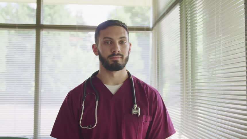 Young bearded male nurse wearing purple scrubs and stethoscope looks at the camera