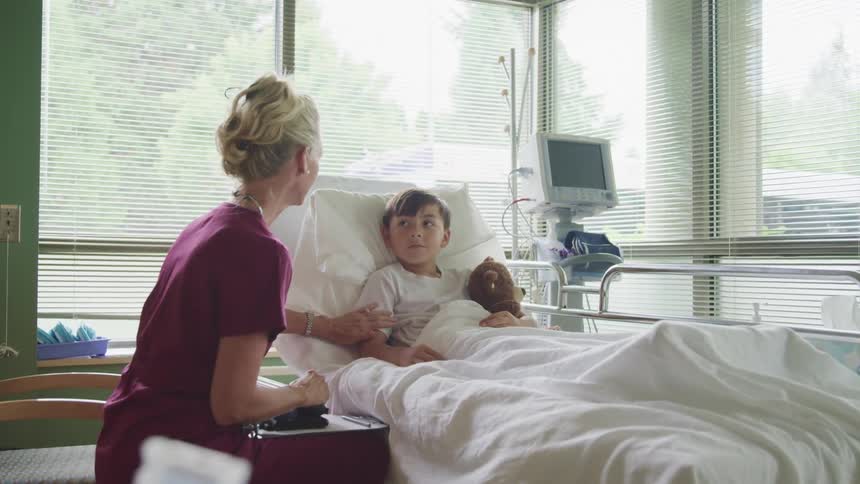 A nurse touches a young male patient on his upper arm as the boy hugs a teddy bear