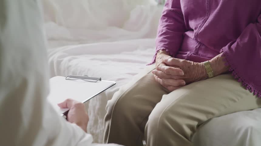 Elderly patient sits on a bed while a doctor stands nearby holding a clipboard