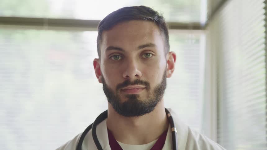 Doctor with a beard and a stethoscope around neck faces the camera with eye contact in the clinic