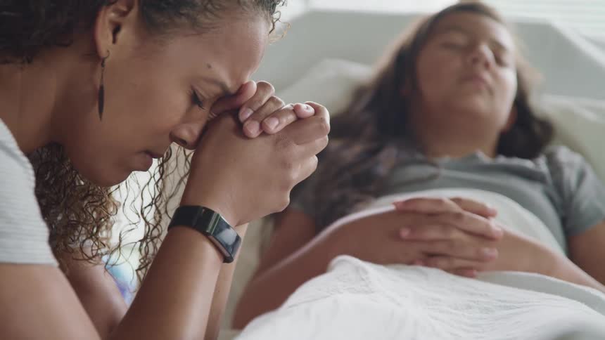 A teenage girl lays in a hospital bed with her mother leaning against the bed