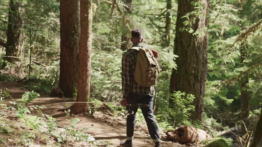 Male carrying a backpack on his back and carrying a fishing pole walks along a hiking trail in the woods on a sunny day