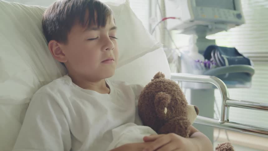 Male child lying in hospital bed holds a teddy bear in his arms as he sleeps