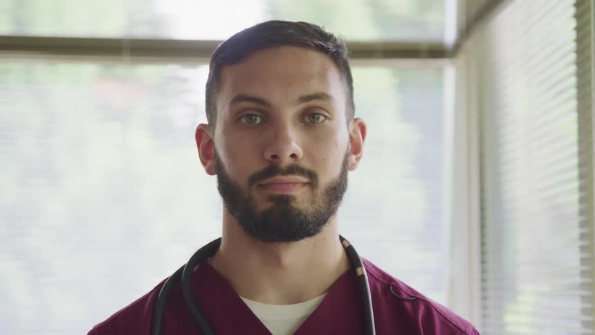 Young male nurse standing in a room near a window looks at the camera