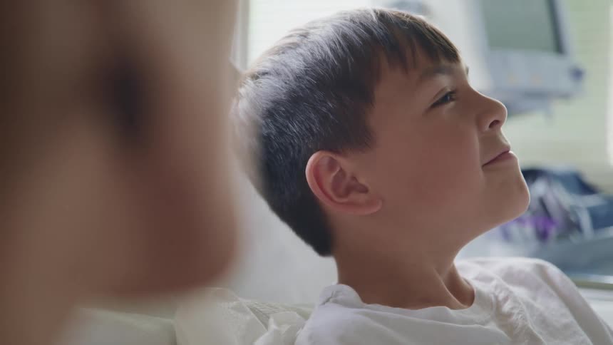 A smiling boy lies in a hospital bed as an adult sits next to the bed