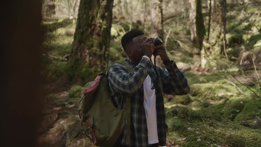 Young man wearing a backpack takes pictures in the forest with a camera at day