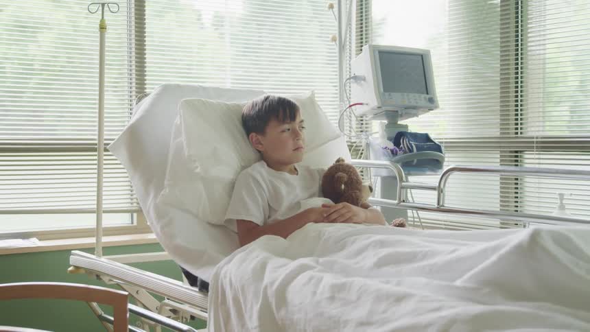 A boy in a hospital bed holding a teddy bear with windows in the background