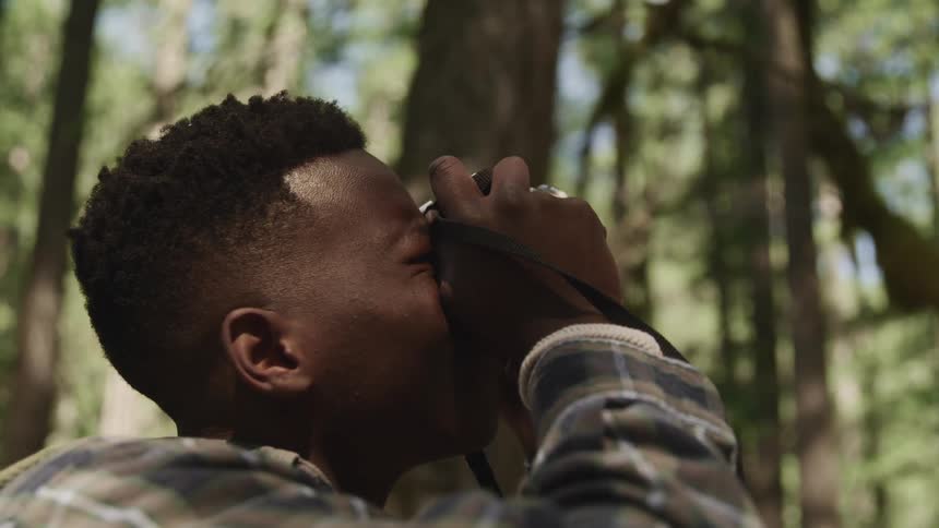 Side view of a black teenage boy taking pictures with a camera in the forest