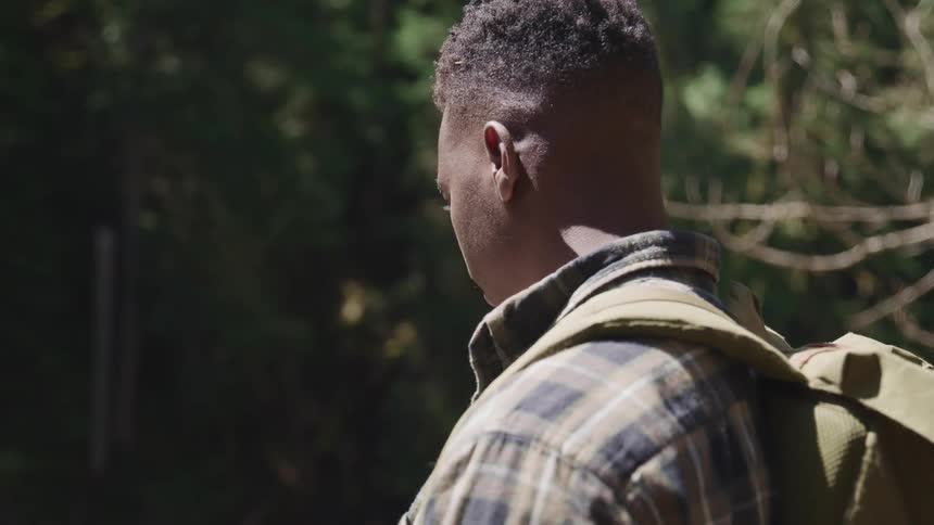 Black man with a backpack and a striped shirt stands and looks down in the forest on a sunny day
