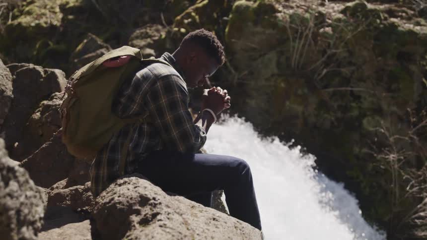 A hiker clasps his hands in prayer as he sits on a rock next to a waterfall