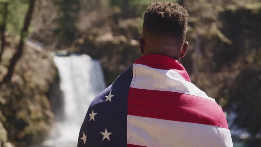 Young black man wrapped in the american flag standing across from a waterfall