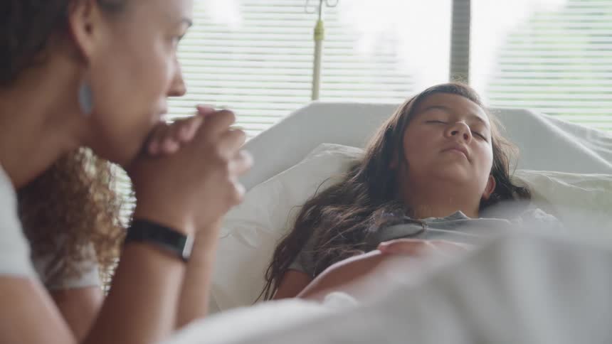 A mother sits beside her daughter laying in a hospital bed looking fearful