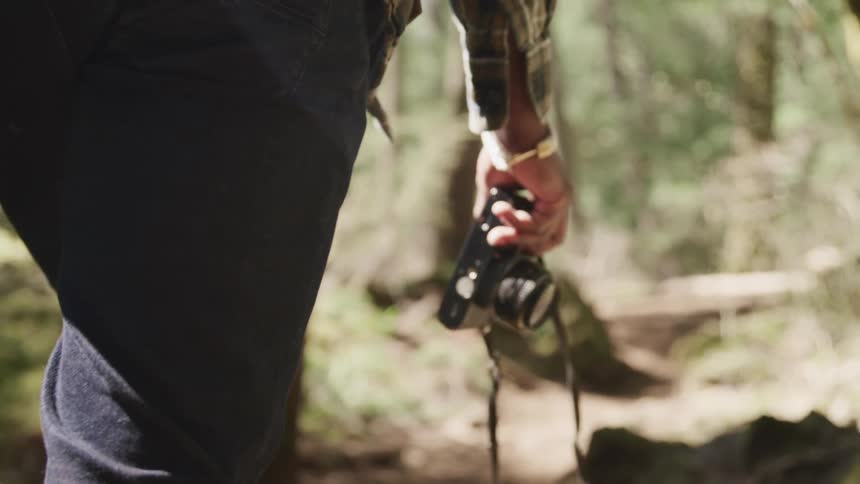 Black man with a shirt and black pants holds a camera and walks on a dirt track in the forest