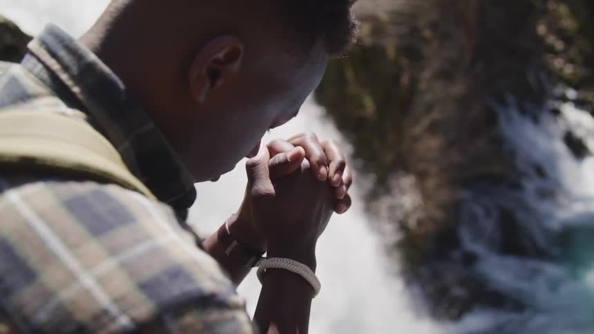 A backpacker bows his head and prays near a waterfall