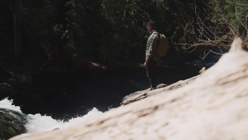 Man wearing a backpack looks at the turbulent waters on a river in the forest