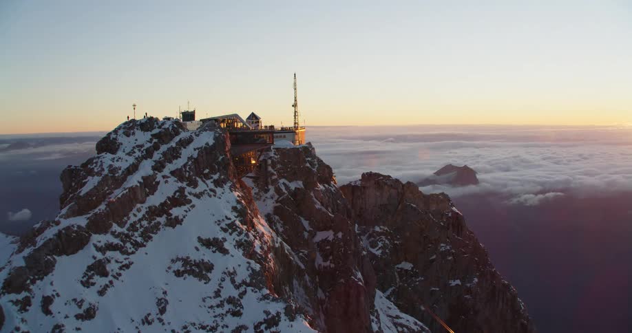 Building occupies peak of snow covered mountain sitting above clouds as sun rises on horizon
