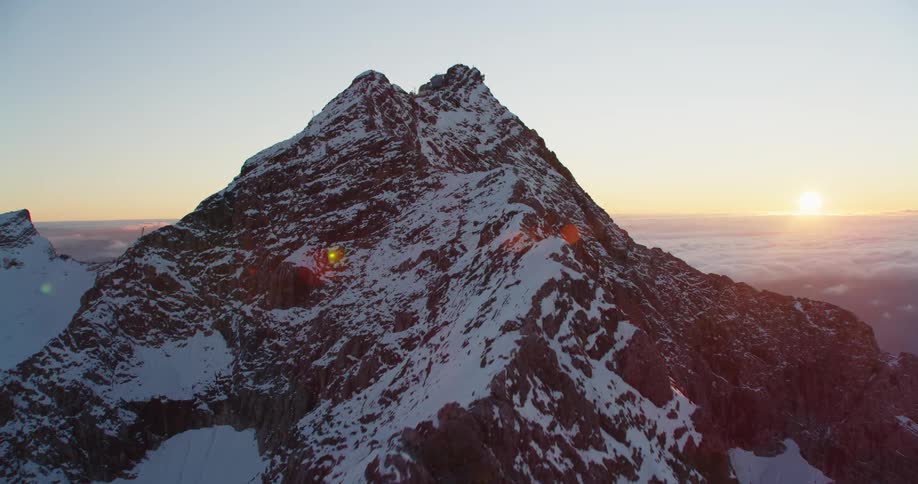 Snow covered mountain towers over surrounding county side as sun sits on the horizon in a red tinted sky